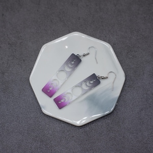 Op de afbeelding: Een paar zilveren oorhangers met een verloop van paars, wit en grijs. De oorhangers hebben een rechthoekige vorm met een uitgesneden ontwerp van halve manen.