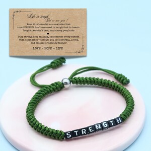 Puede incluir: Pulsera trenzada verde con la palabra "STRENGTH" deletreada en cuentas de letras blancas y negras. La pulsera está sobre una superficie rosa, con una tarjeta que dice "Life is tough, but so are you!" y "LOVE - HOPE - LIFE".