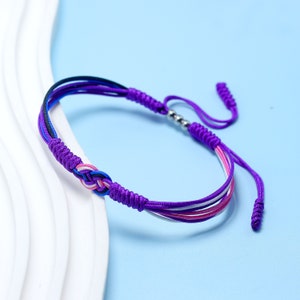 May include: A purple friendship bracelet with a sliding knot fastening. The bracelet has multiple strands in shades of purple, blue, pink, and white. The central knot is intricately woven, and the bracelet is adjustable.