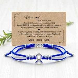 May include: A blue and white braided bracelet with a silver ribbon charm. The bracelet is displayed in front of a card with inspirational text. The card reads "Life is tough, but so are you!"