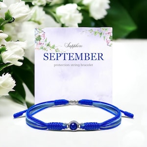 May include: Blue string bracelet with a sapphire gemstone bead. The bracelet is on a white card with the text "Sapphire SEPTEMBER protection string bracelet".