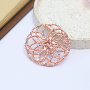 May include: A circular brooch crafted from rose gold-toned twisted wire, featuring an intricate, repeating geometric pattern of interconnected loops that form a flower-like design with a central point where the metal strands are joined together.