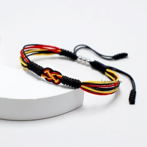 May include: A black, red, and yellow braided bracelet with a knot in the center. The bracelet has a silver bead and a black tassel.