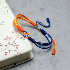 May include: A blue, white, and orange braided friendship bracelet with a silver bead and knotted ends.