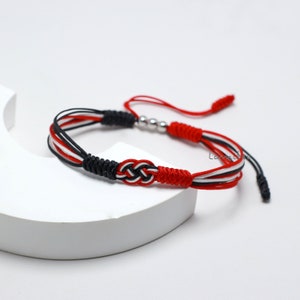 Könnte beinhalten: Rotes, schwarzes und weißes geflochtenes Armband mit einer silbernen Perle. Das Armband hat ein Knotendesign und einen Quasten.