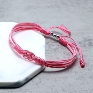 May include: A pink and white braided friendship bracelet with silver beads. The bracelet is tied with a knot and has a long tassel.