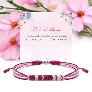 Puede incluir: Pulsera de cuerda rosa, blanca y plateada con una cuenta plateada. La pulsera está sobre una tarjeta blanca con el texto "Badass Mama" y "pulsera de cuerda de protección en código morse" en negro.