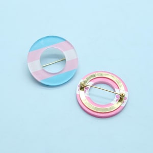 May include: Two round brooches with a pin backing. One brooch features a translucent design with light blue, white, and pink stripes. The other brooch has a pink and white striped design with a gold-colored metal backing.
