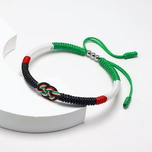 Peut inclure: Un bracelet tressé vert, blanc et noir avec un motif noué au centre. Le bracelet est doté de perles argentées et d'un cordon vert avec un nœud à l'extrémité.