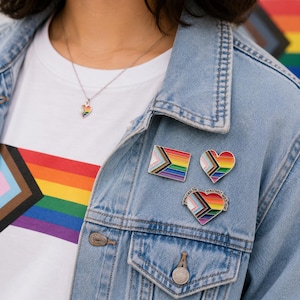 May include: Three enamel pins attached to a light blue denim jacket. One pin is rectangular, one is heart-shaped, and the third is a heart-shaped pin with the text "you can be yourself with me" written along the top edge. All feature the Progress Pride flag.