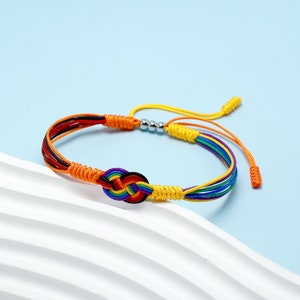 Puede incluir: Una colorida pulsera de la amistad con un diseño de nudo tejido. La pulsera incorpora hebras rojas, negras, naranjas, amarillas, verdes, azules y moradas. La pulsera ajustable tiene un cierre de nudo corredizo y cuentas plateadas.