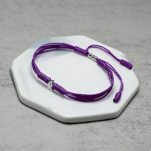 K&ouml;nnte beinhalten: Lilafarbenes geflochtenes Armband mit einem silbernen Schleifenanh&auml;nger und silbernen Perlen.