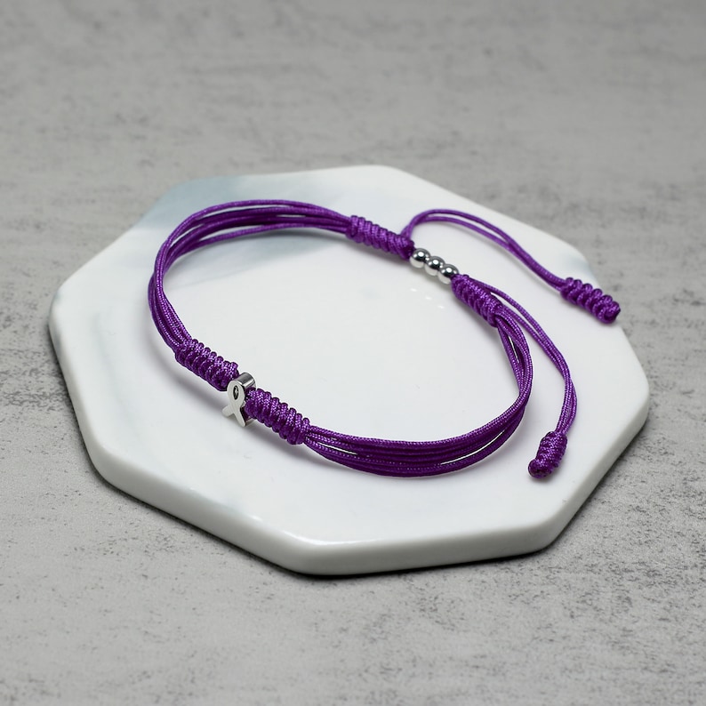 K&ouml;nnte beinhalten: Lilafarbenes geflochtenes Armband mit einem silbernen Schleifenanh&auml;nger und silbernen Perlen.