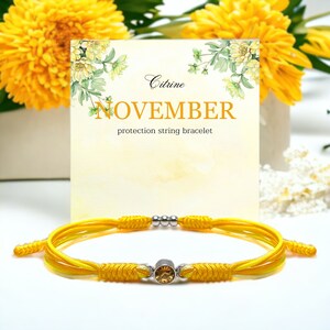 Peut inclure: Bracelet en cordon jaune avec une pierre précieuse en citrine et des perles argentées. Le bracelet est sur un fond blanc avec une carte qui dit "Citrine Novembre bracelet de protection en cordon".