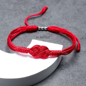 May include: Red string bracelet with a knotted heart design. The bracelet has silver beads and the text #LAMIBEE.