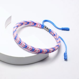 Könnte beinhalten: Ein geflochtenes Freundschaftsarmband in Blau, Rosa und Weiß mit silbernen Perlen. Das Armband hat einen Knotenverschluss.