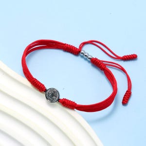 Può includere: Braccialetto rosso regolabile con un medaglione color argento. Il braccialetto è realizzato in cordoncino rosso intrecciato e presenta una chiusura a nodo scorrevole. Il medaglione ha un design dettagliato. Il braccialetto è esposto su una superficie bianca su sfondo azzurro.