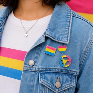 Puede incluir: Una chaqueta de mezclilla azul con tres pines de esmalte: un cuadrado, un corazón y un símbolo de paz, todos con rayas horizontales rosas, amarillas y azules. La chaqueta se lleva sobre una camiseta blanca con rayas horizontales rosas, amarillas y azules.
