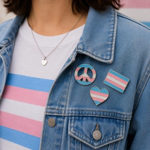 Könnte beinhalten: Drei Emaille-Anstecknadeln an einer blauen Jeansjacke. Die Anstecknadeln zeigen die Farben der Transgender-Pride-Flagge in Hellblau, Rosa und Weiß in Form eines Friedenszeichens, eines Herzens und einer rechteckigen Flagge. Eine Herz-Halskette ist sichtbar.