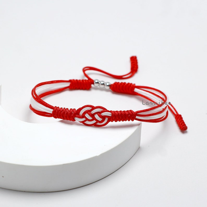 May include: Red and white braided bracelet with a silver bead and a knot design. The bracelet is tied with a knot at each end.