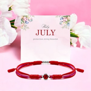 May include: Red, pink and white string bracelet with a red gemstone and silver beads. The bracelet is on a pink background with a card that says "Ruby JULY protection string bracelet".