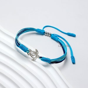 Puede incluir: Un brazalete de cordón trenzado azul y verde con un dije de tortuga plateado. El brazalete tiene un cierre de nudo deslizante.