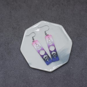 Op de afbeelding: Een paar hangende oorbellen met een verloop van roze, paars en blauw. De oorbellen hebben een uitsnijding van een wassende maan in verschillende fasen.