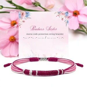Pode incluir: Um bracelete de amizade trançado rosa, branco e bordô com contas prateadas. O bracelete está em um cartão branco com o texto "Badass Sister" e "pulseira de corda de proteção de código morse".
