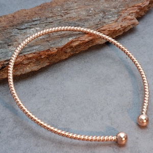 Könnte beinhalten: Ein Armreif aus roségoldfarbenem Metall mit einer gedrehten, seilartigen Textur entlang des gesamten Bandes. Das offene Design endet an beiden Seiten mit einer glatten, polierten kugelförmigen Perle, die den Kreis vervollständigt.