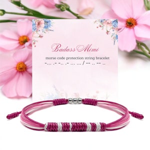 May include: Pink, white, and silver braided string bracelet with silver beads. The bracelet is on a white card with the text "Badass Mimi" and "morse code protection string bracelet" in black text.