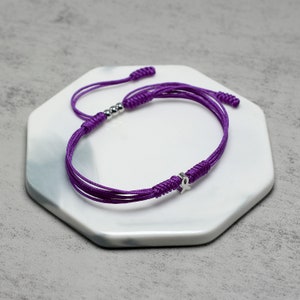 K&ouml;nnte beinhalten: Lilafarbenes Armband aus Schnur mit einem silbernen Band-Anh&auml;nger. Das Armband ist mit einem Knoten gebunden und hat silberne Perlen.