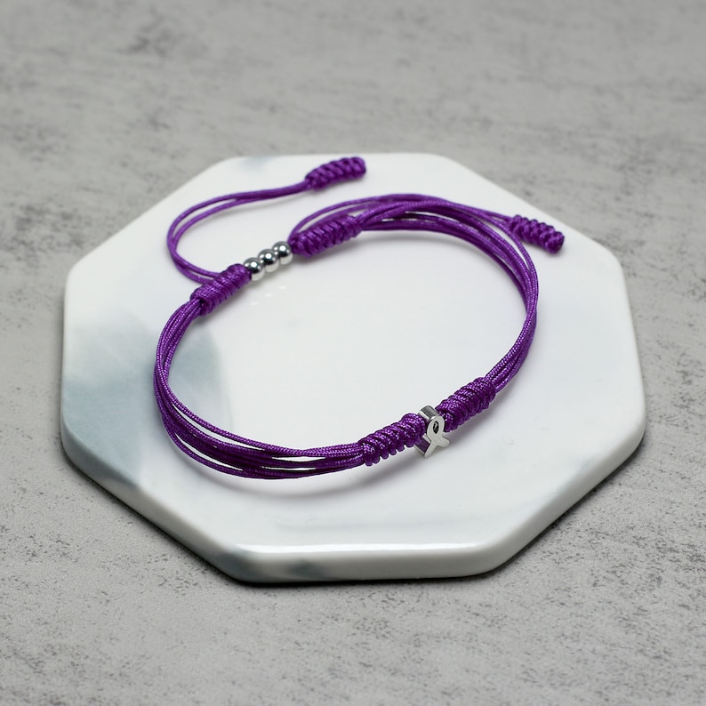 K&ouml;nnte beinhalten: Lilafarbenes Armband aus Schnur mit einem silbernen Band-Anh&auml;nger. Das Armband ist mit einem Knoten gebunden und hat silberne Perlen.