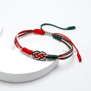 Puede incluir: Un brazalete de amistad trenzado rojo, blanco y verde con un acento de cuentas plateadas. El brazalete está atado en un nudo.