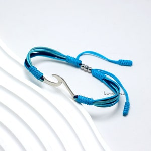 Puede incluir: Un brazalete de cordón trenzado azul y verde con un dije de ola plateado y cuentas plateadas. El brazalete tiene cierres ajustables.