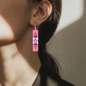 May include: Pink rectangular earrings with a floral and crescent moon design. The earrings have a silver hook and a white stripe in the middle. The flower is purple, and the crescent moons are black.