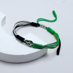 May include: A green, black, and white braided bracelet with a silver bead accent. The bracelet is tied with a knot at the end.