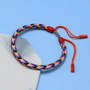 Puede incluir: Pulsera trenzada arcoíris con detalles rojos y cordones naranjas ajustables. La pulsera presenta un diseño tejido con colores como rojo, naranja, amarillo, verde, azul y morado. Sobre un fondo azul.