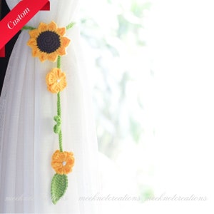 May include: A crocheted sunflower curtain tieback with yellow and green flowers and leaves. The tieback is hanging on a white curtain.