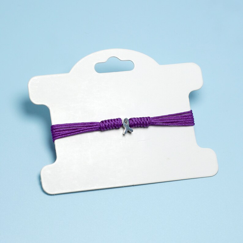 K&ouml;nnte beinhalten: Ein Armband aus lila Kordel mit einem silbernen Schleifenanh&auml;nger. Das Armband wird auf einer wei&szlig;en Karte mit einem Loch zum Aufh&auml;ngen pr&auml;sentiert.
