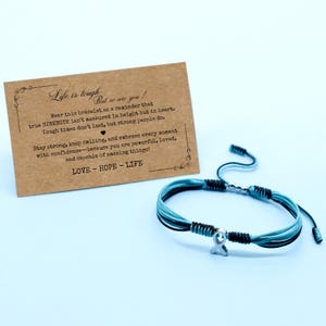 May include: A multi-strand bracelet with blue, black, and silver accents. The bracelet features a silver ribbon charm and an adjustable closure. A card with the text "Life is tough, but so are you!" is in the background. The card also includes the words "LOVE - HOPE - LIFE".