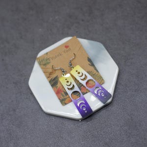 May include: A pair of dangle earrings with a yellow to purple ombre gradient. The earrings feature a rectangular shape with a cutout design of crescent moons.