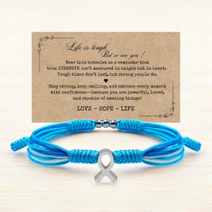 May include: A blue and white cord bracelet with a silver ribbon charm. The bracelet is displayed in front of a card with the text "Life is tough, but so are you!" and "LOVE - HOPE - LIFE".