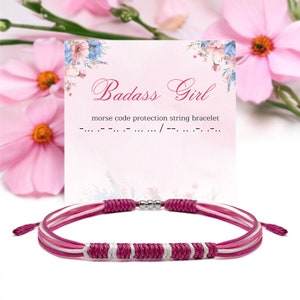 Op de afbeelding: Een roze en witte touw armband met een magnetische sluiting. De armband wordt getoond met een kaart met de tekst "Badass Girl" en "Morsecode bescherming touw armband". De achtergrond heeft roze bloemen.
