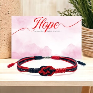 May include: Red and black string bracelet with a knot in the center. The bracelet is on a wooden surface and a card with the word "Hope" and the text "protection string bracelet" is behind it.