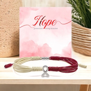 May include: A red and cream colored string bracelet with a silver ribbon charm. The bracelet is on a pink and white background with the word "Hope" written in red.