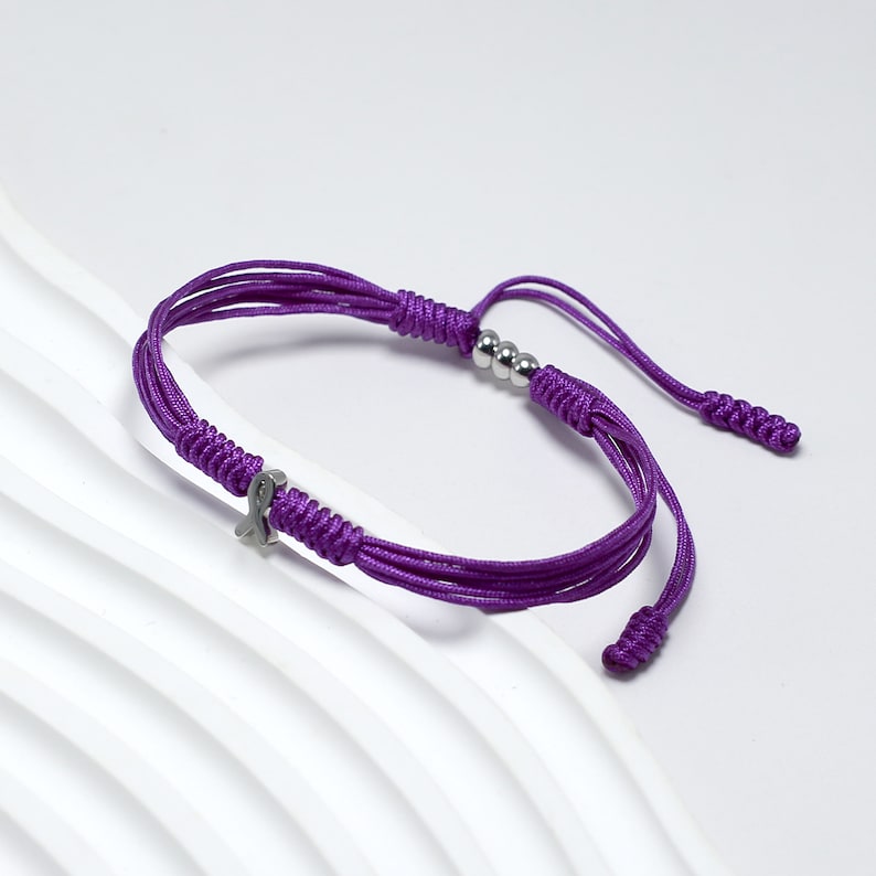 K&ouml;nnte beinhalten: Lilafarbenes geflochtenes Armband mit einem silbernen Schleifenanh&auml;nger und silbernen Perlen.