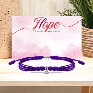 K&ouml;nnte beinhalten: Lilafarbenes Armband aus Schnur mit einem silbernen Band-Anh&auml;nger. Das Armband liegt auf einer Holzoberfl&auml;che mit einer rosa Karte, auf der "Hope protection string bracelet" steht.