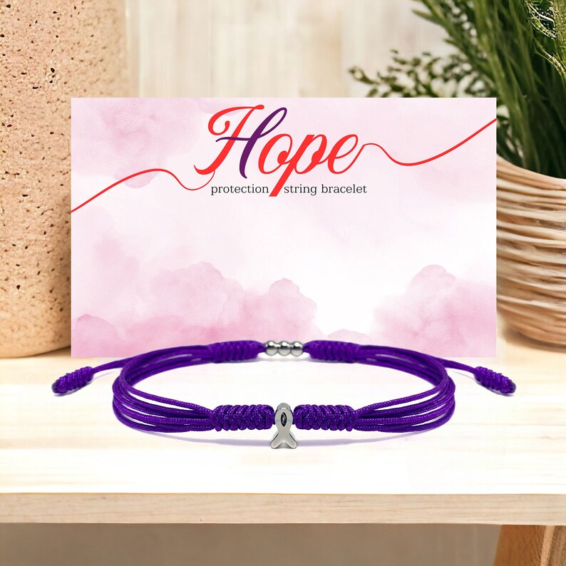 K&ouml;nnte beinhalten: Lilafarbenes Armband aus Schnur mit einem silbernen Band-Anh&auml;nger. Das Armband liegt auf einer Holzoberfl&auml;che mit einer rosa Karte, auf der "Hope protection string bracelet" steht.