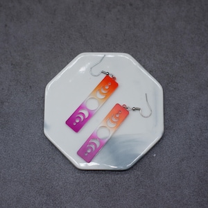 Könnte beinhalten: Ein Paar Hängeohrringe mit einem Farbverlauf von Pink zu Orange. Die Ohrringe haben ein ausgeschnittenes Design mit einem Halbmond und Vollmondphasen.