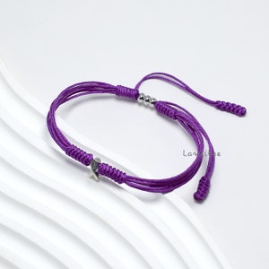 K&ouml;nnte beinhalten: Lilafarbenes Armband aus Schnur mit einem silbernen Band-Anh&auml;nger und silbernen Perlen. Das Armband ist an beiden Enden mit einem Knoten befestigt.